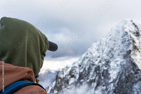 Fototapeta man hiker wearing a green hoodie and cap, gazes at a snow-covered mountain range under a cloudy sky, winter exploration
