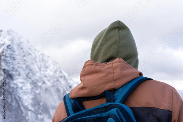 Fototapeta person with a green hood and brown jacket stands against a snowy mountain backdrop. The scene captures a sense of adventure in a winter landscape, hiking