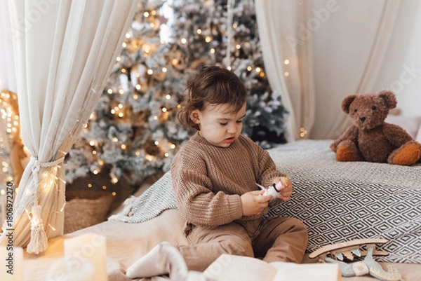 Fototapeta Little boy sitting on the bed in front of christmas tree. Toddler playing with toys.