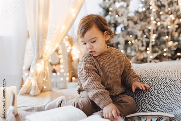 Fototapeta Little boy sitting on the bed in front of christmas tree. Toddler playing with toys and candles. Brown colors.