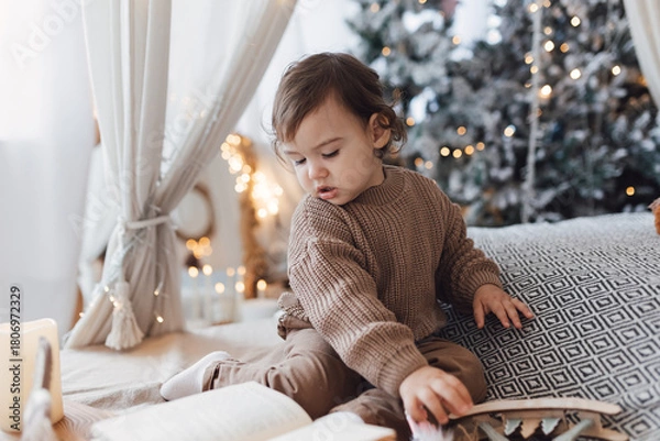 Fototapeta Little boy sitting on the bed in front of christmas tree. Toddler playing with toys and candles. Brown colors.