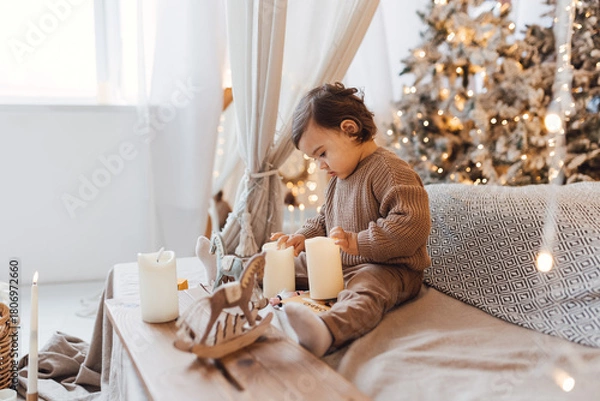 Fototapeta Little boy sitting on the bed in front of christmas tree. Toddler playing with toys and candles. Brown colors.