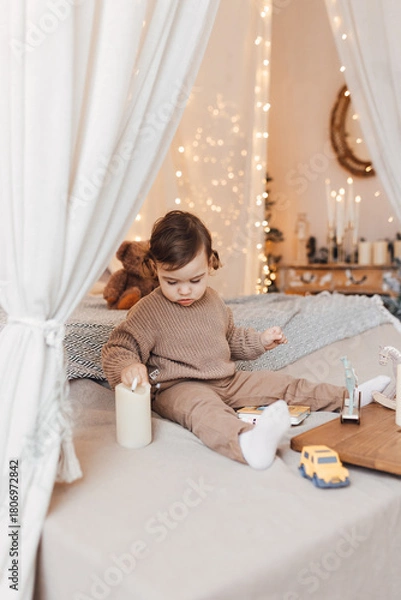 Fototapeta Little boy sitting on the bed in front of christmas tree. Toddler playing with toys and candles. Brown colors.