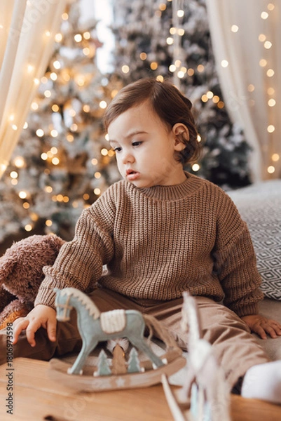 Fototapeta Little boy sitting on the bed in front of Christmas tree. Toddler playing with toys