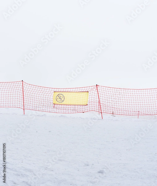 Fototapeta A snowy landscape with a red safety net and a warning sign. The scene is foggy, creating a sense of isolation and caution in winter conditions.