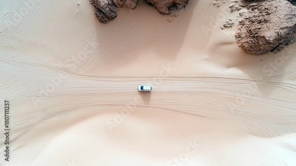 Fototapeta An aerial perspective captures a solitary white vehicle traversing a vast, sandy desert landscape, leaving tire tracks on the smooth dunes.