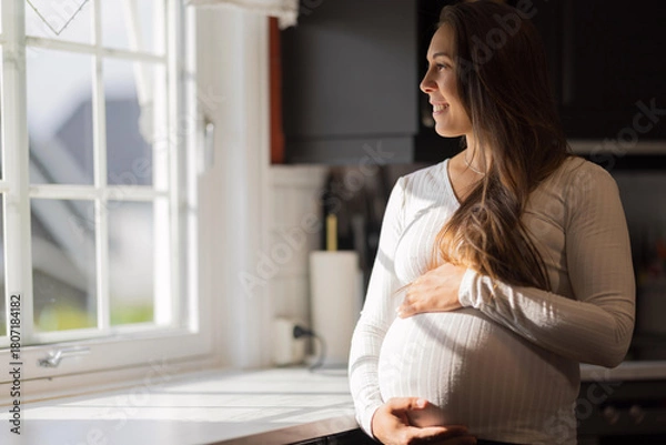 Fototapeta Smiling pregnant woman standing by a window in bright sunlight
