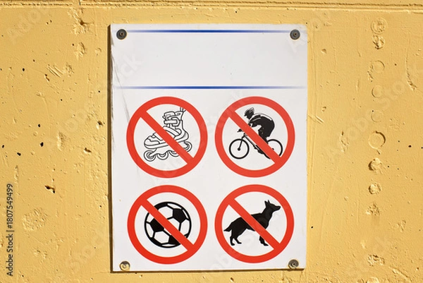 Obraz A white informational panel with red prohibition signs for rollerblades, bicycles, ball games, and dog walking, mounted on a bright yellow wall in a public space.