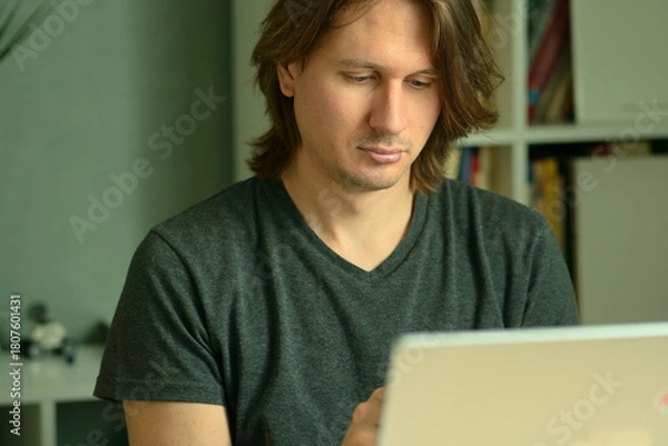 Fototapeta A young man with long hairstyle working on his laptop at home office desk. Focused look at the computer screen. Remote work and telecommuting concept.