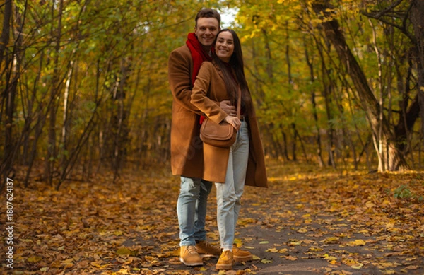 Fototapeta Smiling couple standing and hugging in autumn park, dressed in matching brown coats and jeans, surrounded by colorful yellow leaves and warm fall atmosphere.