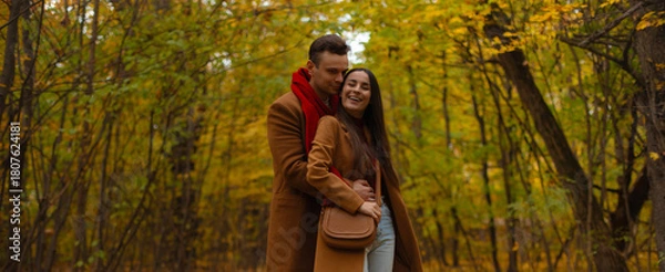 Fototapeta Smiling couple embracing in autumn forest, dressed in brown coats and red accents, enjoying romantic walk surrounded by colorful fall foliage and golden leaves.