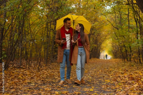Fototapeta Happy couple walking under yellow umbrella in autumn park, dressed in matching brown coats and red accents, smiling and enjoying romantic walk surrounded by colorful fall foliage.