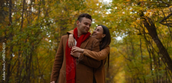 Obraz Happy couple in brown coats embracing in autumn park, smiling and looking at each other with love, surrounded by golden foliage and warm seasonal colors of fall.
