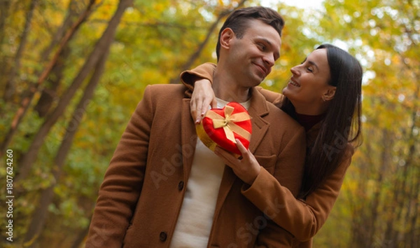 Fototapeta Romantic couple celebrating Valentine’s Day in an autumn forest. Woman hugs her partner and gives him a heart-shaped gift box, sharing a warm and loving moment.
