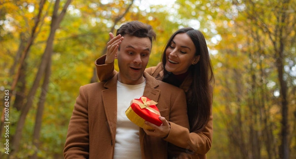 Obraz Happy couple celebrating Valentine’s Day in an autumn forest. Woman surprises her partner with a heart-shaped gift box, and he reacts with joy and excitement.
