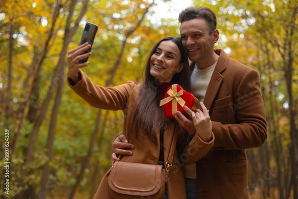 Fototapeta Happy couple taking a selfie on Valentine’s Day in an autumn forest. Woman holds a heart-shaped gift box while they smile and enjoy a romantic moment together.
