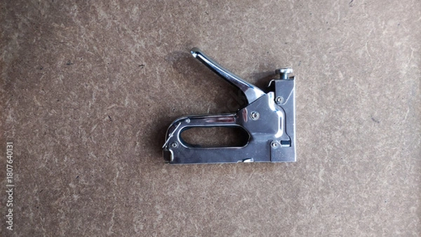 Fototapeta Top view of shiny metal staple gun placed on a brown textured surface, showcasing a common hand tool used for fastening materials.