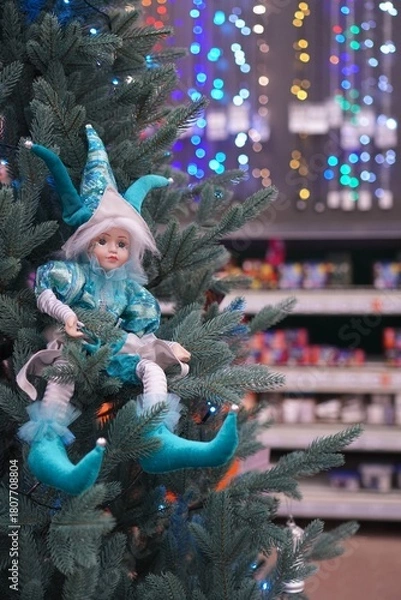 Fototapeta Vintage elf figurine in blue suit on artificial Christmas tree on the background of shelves with toys and colorful garlands out of focus as a new year gift shopping concept