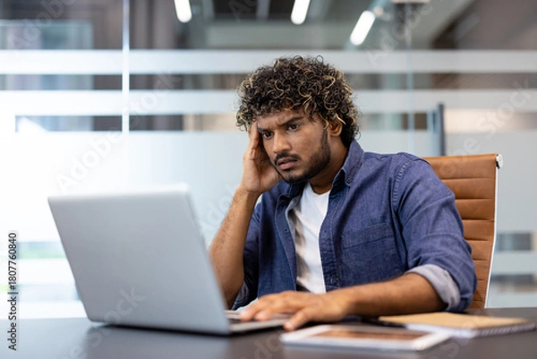Fototapeta Upset young Indian man sitting at a desk in the office and looking at the laptop screen in frustration
