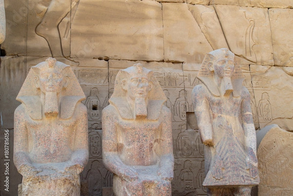 Fototapeta Egypt, Luxor, 01.02.2022.Ancient Egyptian stone statues standing and seated among sandstone temple ruins with carved hieroglyphs and warm light, Karnak temple.