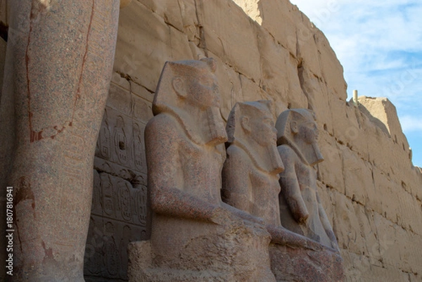 Fototapeta Egypt, Luxor, 01.02.2022.Ancient Egyptian stone statues standing and seated among sandstone temple ruins with carved hieroglyphs and warm light, Karnak temple.