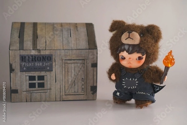 Obraz Turkey, October 19, 2025: a figurine of a fairy-tale character, a Korean boy dressed as a bear holding a flaming torch, next to a cardboard packaging box on a gray background