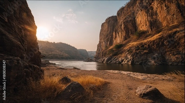 Fototapeta Bright sun sets behind tall rocky cliffs that frame a calm river. The warm light reflects off the water while dry grass and stones line the sandy shore in the fading day.