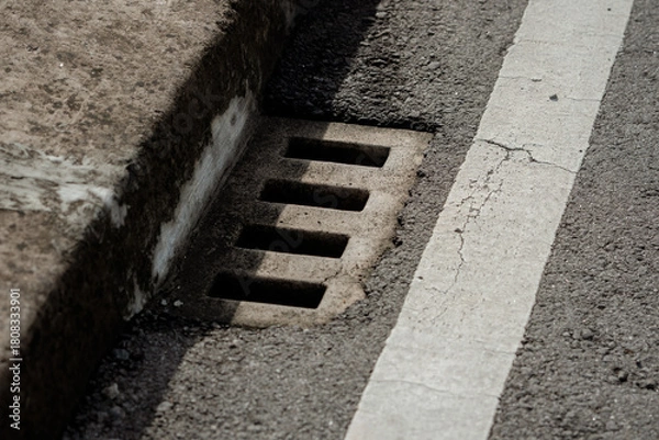 Fototapeta Concrete drainage grate on an asphalt road side next to a white painted traffic line markings.