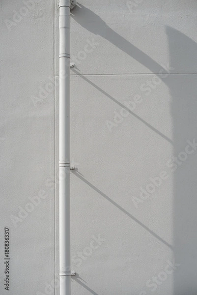 Fototapeta Vertical white drainage pipe casting a long diagonal shadow on a textured gray concrete wall surface.