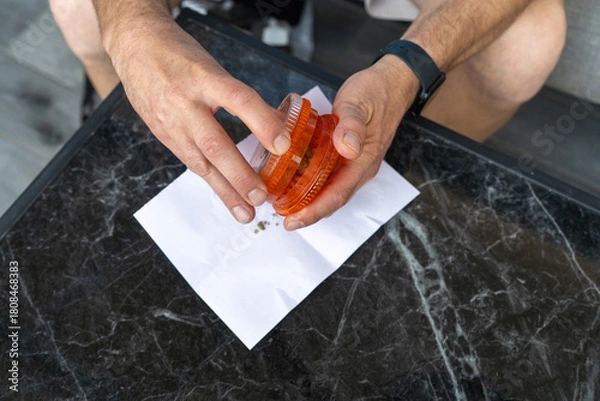 Fototapeta Close-up of a middle-aged mans hands using an herb grinder to prepare cannabis on a marble table, highlighting a therapeutic ritual that blends recreational and medicinal cannabis use. 