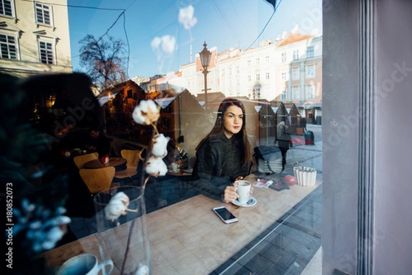 Fototapeta Beautiful brunette young woman in a cafe holding a cup of coffee or cocoa, seen through the window with buildings and lights reflections. She is looking away. Lifestyle concept.