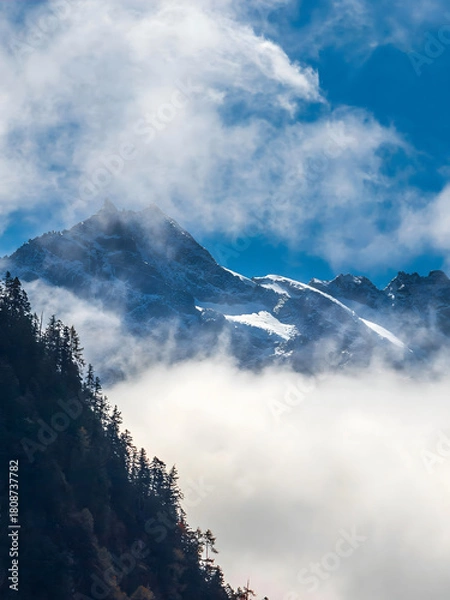 Obraz Snowcapped Mountain Peaks Through Misty Clouds with Forested Slopes