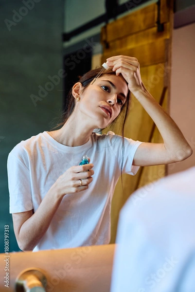 Fototapeta Young woman applies a cosmetic serum to her forehead with a dropper in front of a mirror, performing a morning routine