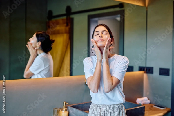 Obraz Woman standing in bathroom, gently massaging moisturizer into her face with her eyes closed, practicing self-care