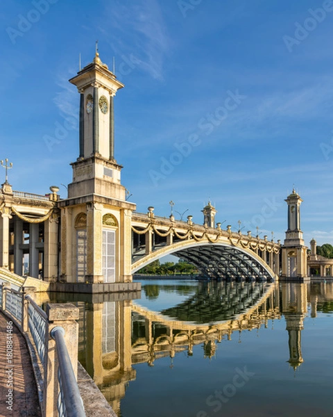 Obraz Grand Seri Gemilang Bridge with ornate towers and clock faces, reflected in calm water under a blue sky in Putrajaya, Malaysia