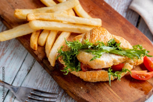 Fototapeta chicken croissant sandwich served with a side of french fries on a wooden board. The sandwich is made with a flaky croissant and filled with a piece of chicken, green lettuce, and sliced red tomatoes.