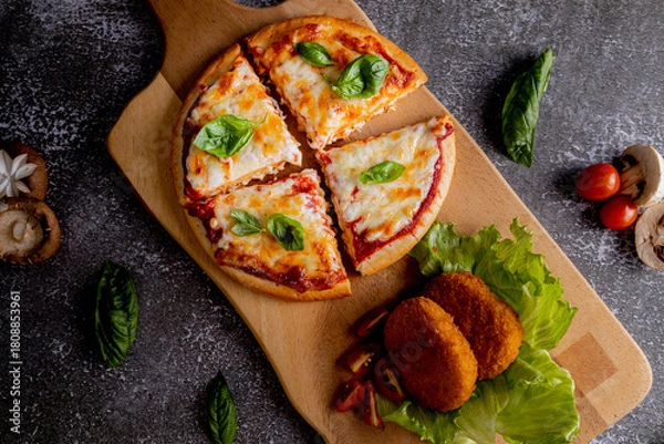 Fototapeta Vegetarian pizza cut into four slices, topped with melted cheese, tomato sauce, and fresh basil leaves. Accompanying the pizza are two breaded, oval-shaped croquettes resting on a bed of lettuce