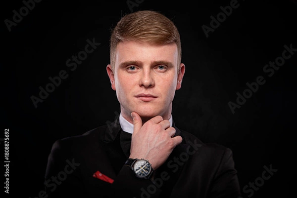 Fototapeta A handsome young Caucasian man in a black tuxedo and bow tie, wearing a wristwatch, holds his chin while looking directly at the camera against a dramatic black background.