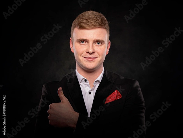 Fototapeta Studio portrait of a smiling young businessman with red hair wearing a black suit and showing a thumbs up gesture against a dark background.