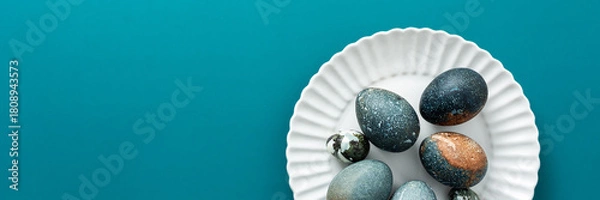 Fototapeta A collection of intricately decorated Easter eggs in various shades rests on a white plate against a soothing teal backdrop, perfect for spring festivities and family traditions, banner