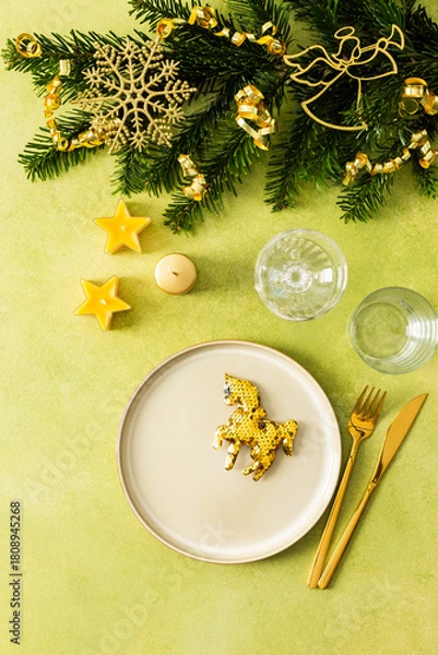 Fototapeta A festive Christmas table setting with an empty plate and cutlery on a bright green background. Spruse branches and golden tree decorations create a warm, holiday atmosphere