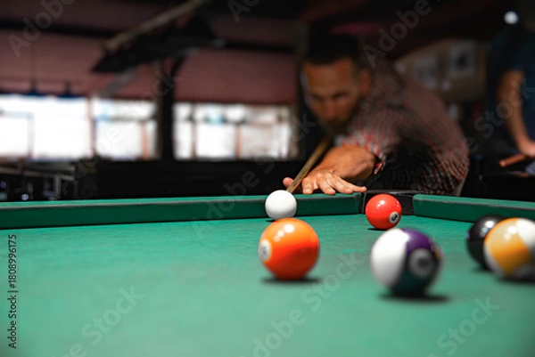 Fototapeta Close-up view of colorful billiard balls on a green felt table with a blurred male player aiming his cue stick in the background.