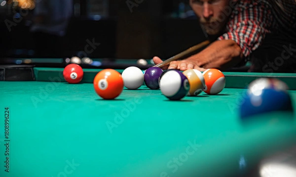 Fototapeta A close-up view of colorful billiard balls scattered on a green felt table while a focused man in a plaid shirt aims his cue stick in the blurred background.