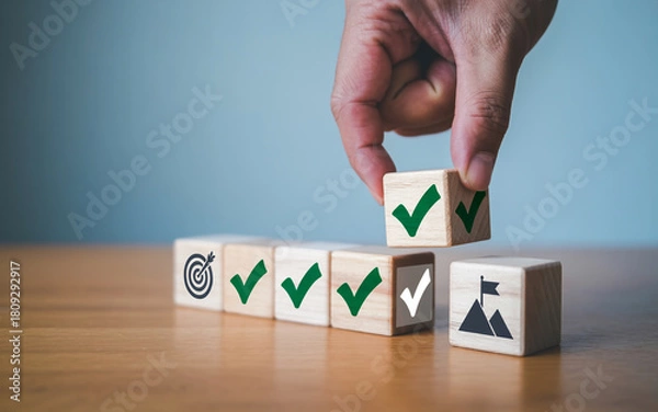 Fototapeta Hand placing a wooden block with a checkmark to complete a sequence representing goal achievement and project success on a wooden table against a blue background