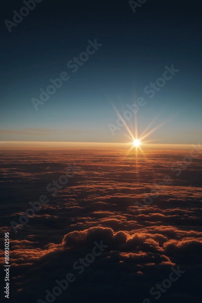 Obraz Golden-hour sunrise above sculpted atmospheric cloud layers, warm inner glow contrasting with cool blue shadows, intense central starburst flare