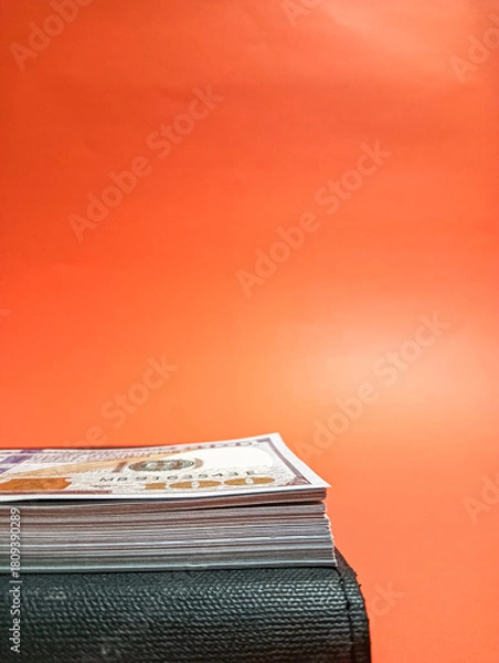 Fototapeta A neat stack of cash placed on a black ledger with a bold orange backdrop, representing wealth, financial planning, budgeting, and economic concepts in a clean minimal composition.