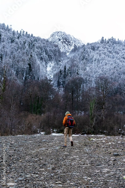 Fototapeta young man hiker with a backpack walks on a rocky path in a snowy mountain landscape. Trees are covered in frost, and mountains rise in the background