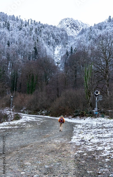 Fototapeta person hiker with backpack walks along a snowy path in a mountainous area. The landscape features bare trees and snow-covered ground, creating a serene winter scene