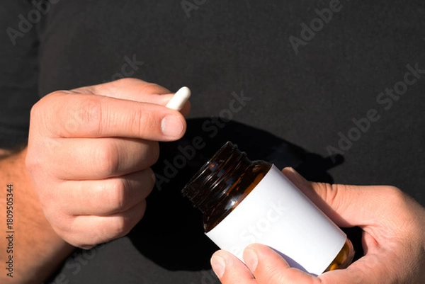 Fototapeta Hands present a capsule above an amber bottle with white label against dark fabric; bright sunlight, clean lines and negative space convey vitamin health and confident lifestyle.