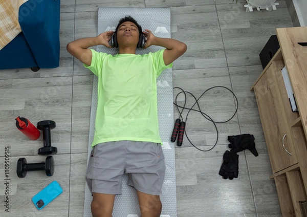 Obraz Top view of a young man lying on the exercise mat with his eyes closed, listening to music and resting after working out. A moment of relaxation and well being